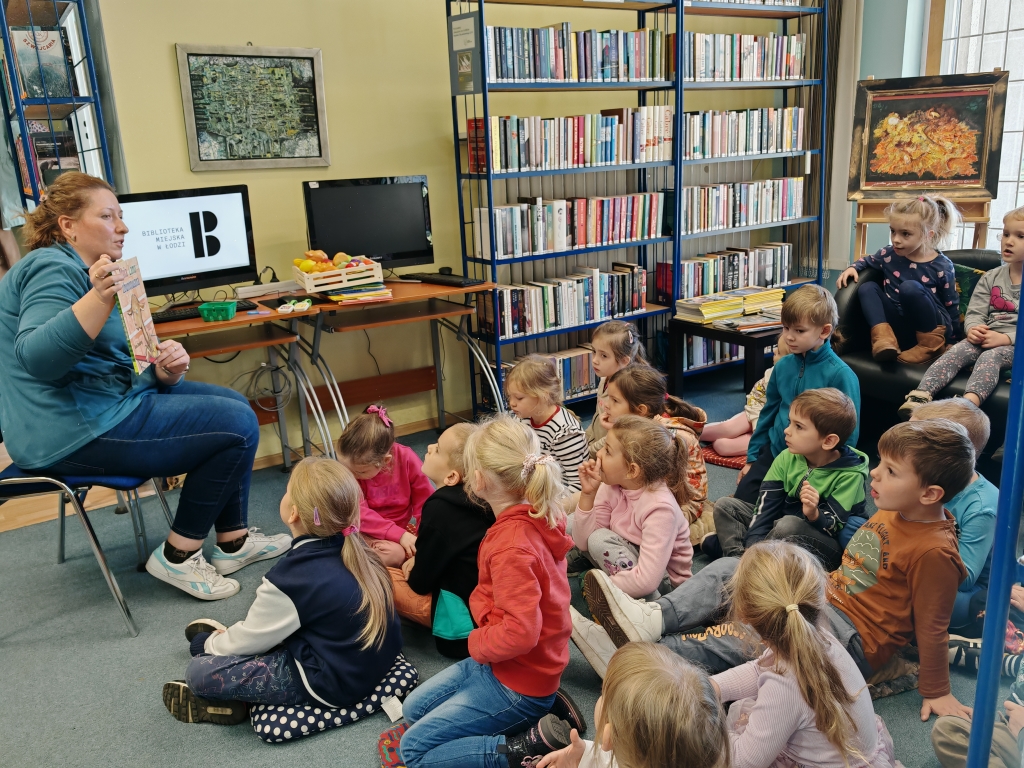 Zajęcia w Bibliotece - dzieci siedzą na dywanie i słuchają prowadzącej